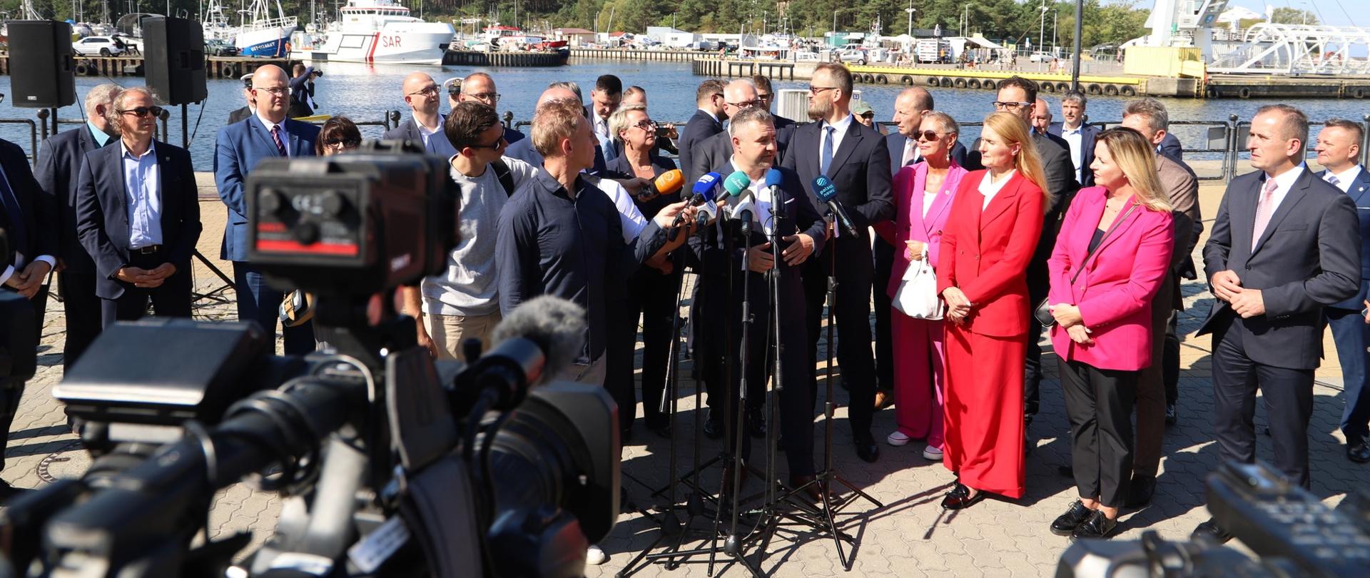 grupa osób stoi na tle portu w Ustce, przed nimi mikrofony