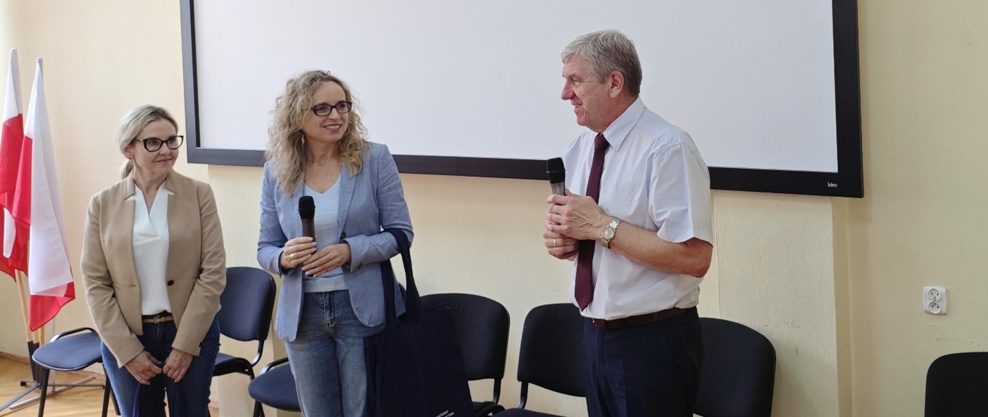 Trzy osoby rozmawiają w sali konferencyjnej – dwie kobiety i jeden mężczyzna. Jedna z kobiet i mężczyzna trzymają mikrofony. W tle widać biały ekran projekcyjny, kilka krzeseł oraz flagi Polski.