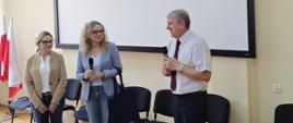 Trzy osoby rozmawiają w sali konferencyjnej – dwie kobiety i jeden mężczyzna. Jedna z kobiet i mężczyzna trzymają mikrofony. W tle widać biały ekran projekcyjny, kilka krzeseł oraz flagi Polski.