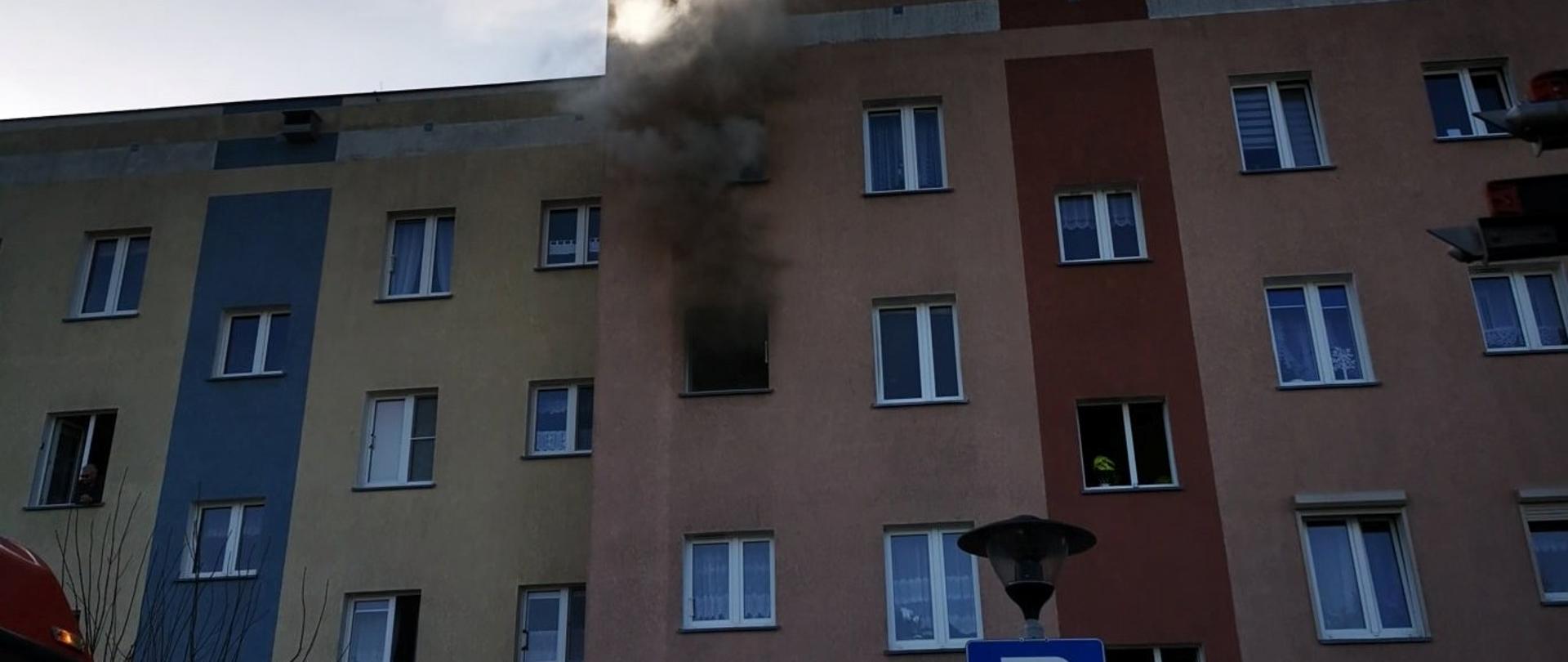 Widać budynek mieszkalny (blok) - z okna na drugim piętrze wydobywa się dym