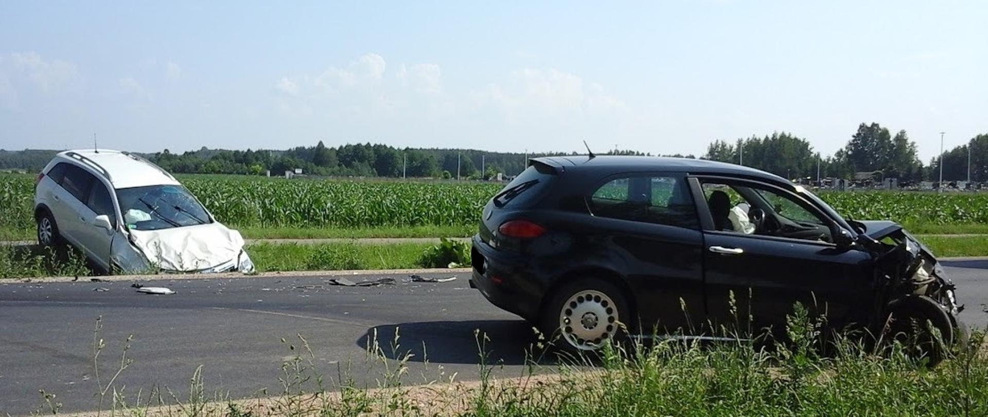 Uszkodzone pojazdy po zderzeniu. W obu uszkodzone są przednie części pojazdów. Jeden stoi na jezdni, drugi na poboczu , przodem w rowie.