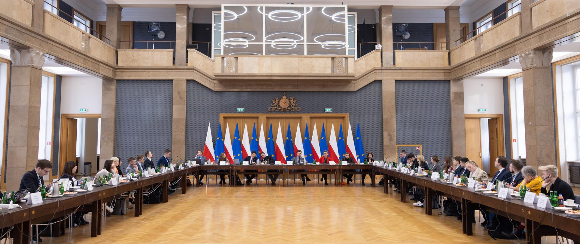 Szeroki kadr. Uczestnicy posiedzenia Rady obradują w sali konferencyjnej przy stole ustawionym w literę U. W tle widoczne flagi Polski i Unii Europejskiej, po bokach widać sali kolumny i okna.