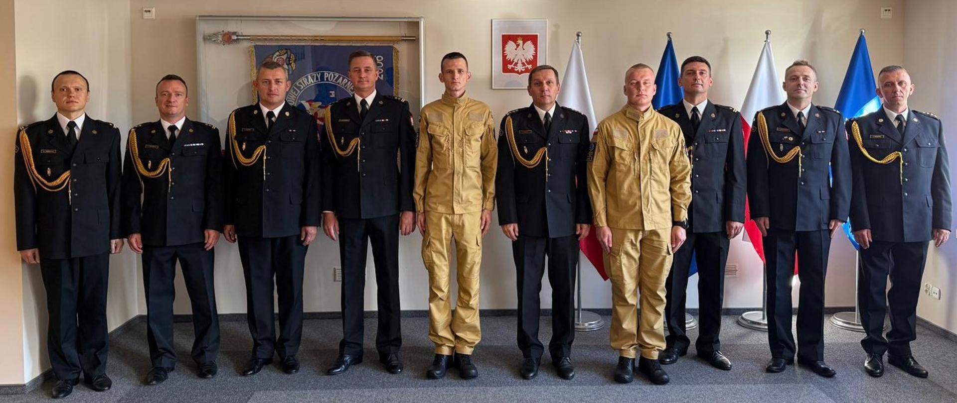 Fotografia przedstawia grupę dziesięciu strażaków ustawionych w jednym rzędzie w oficjalnym otoczeniu. Ośmiu z nich ma na sobie galowe, ciemnogranatowe mundury ze złotymi dystynkcjami, sznurami galowymi i medalami. Dwaj strażacy, stojący w środkowej części rzędu, wyróżniają się piaskowymi mundurami.