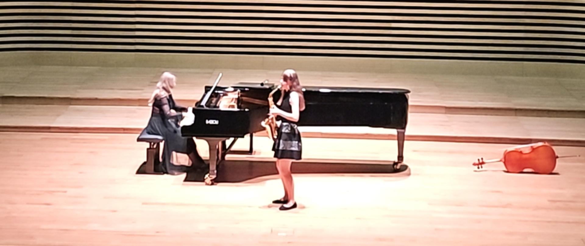 Nastolatka stojąc na estradzie sali koncertowej gra na saksofonie, za nią kobita gra na fortepianie. Z przodu widać od tyłu fragment widowni.