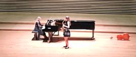 Nastolatka stojąc na estradzie sali koncertowej gra na saksofonie, za nią kobita gra na fortepianie