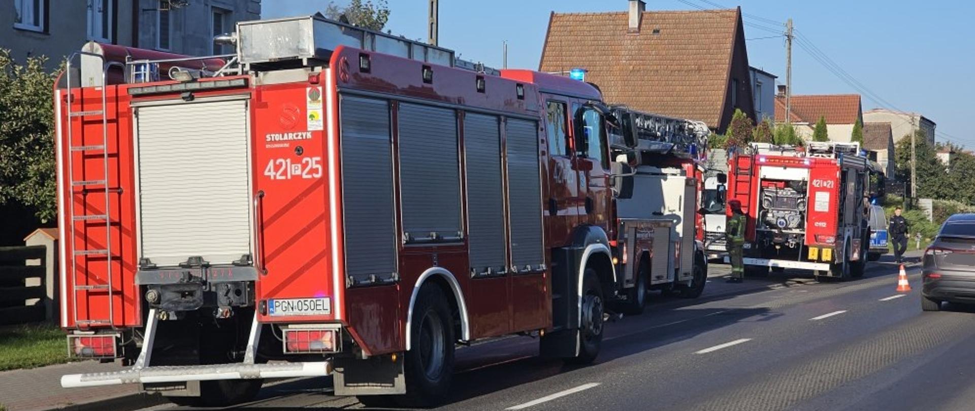 Na zdjęciu widać samochody pożarnicze biorące udział w akcji. Pojazdy stoją na drodze publicznej przed posesją w której powstał pożar.
