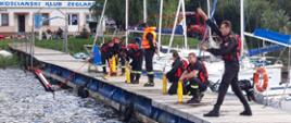 Szkolenia z ratownictwa na obszarach wodnych. Grupa strażaków ubrana w skafandry nurkowe i kamizelki ratunkowe stoi na pomoście i ćwiczy wyrzucanie i zwijanie rzutki ratowniczej. W tle jezioro i drzewa.