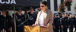 Wojewoda Opolski Monika Jurek w trakcie przemówienia podczas Święta Policji. Stoi przy mównicy, w tle widać pozostałych uczestników wydarzenia. 