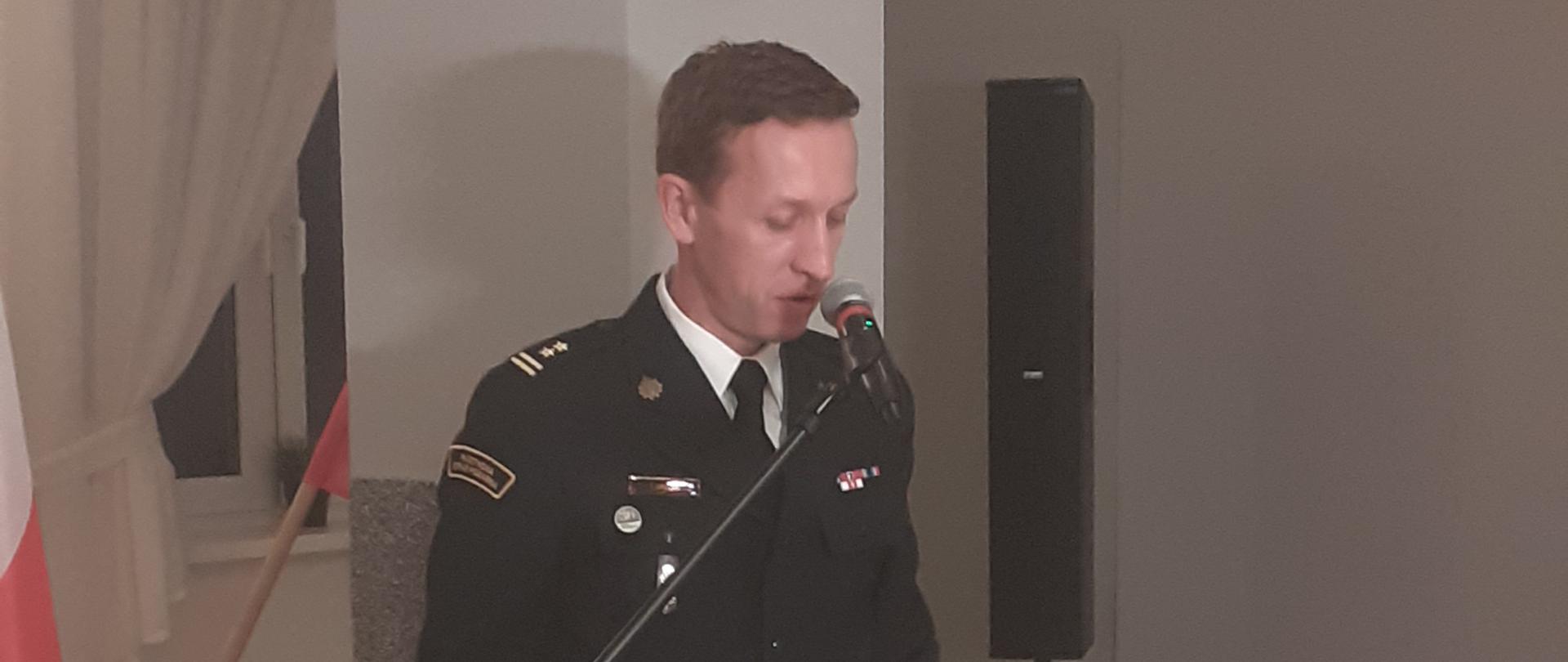 strażak w mundurze mwyjściowym stoi przy mównicy, w tle biały słup i flaga polski