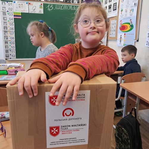 W klasie w ławkach siedzą dzieci z niepełnosprawnością; na pierwszym planie dziewczynka trzyma ręce na paczce z pomocą materialną