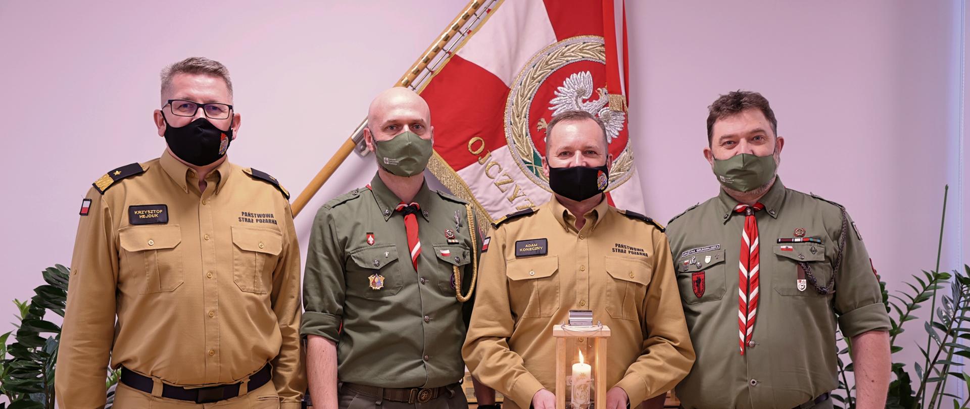 Zastępcy komendanta głównego PSP wraz z druhami pozują do zdjęcia stając naprzemiennie od lewej tj. zastępca komendanta oraz druh, jeden z zastępców trzyma lampion, w tle sztandar na ścianie