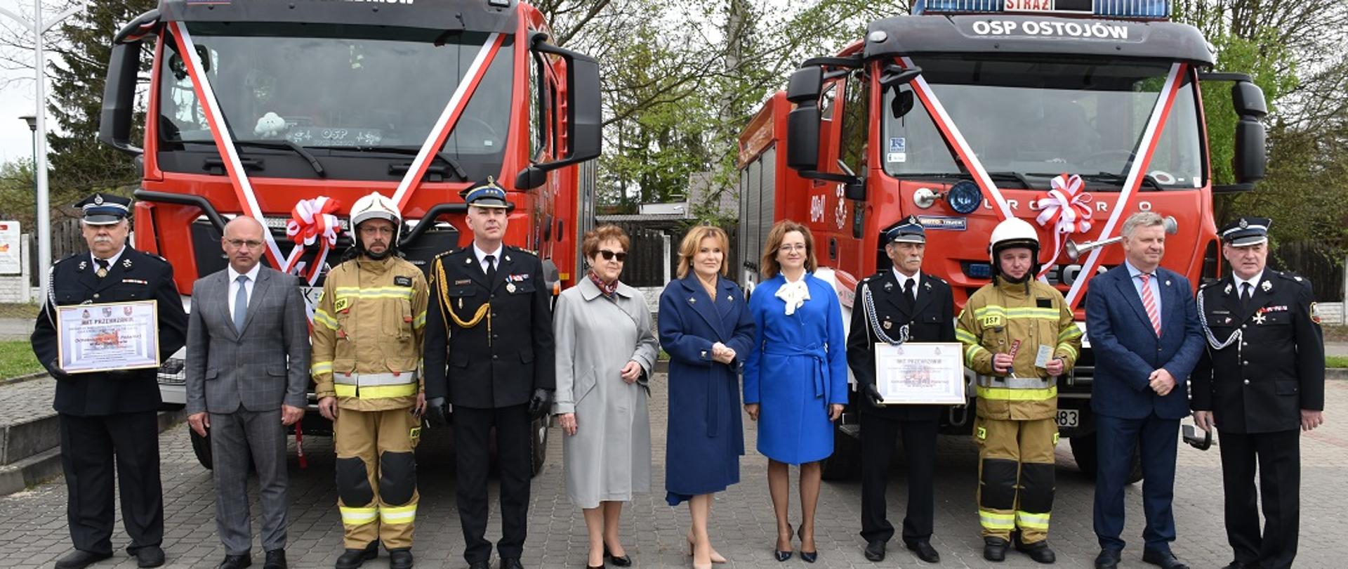 Na zdjęciu na tle dwóch pojazdów gaśniczych widzimy stojące osoby. Część osób ubranych jest w galowe mundury strażackie pozostałe osoby ubrane są po cywilnemu. Wśród stojących osób dwóch strażaków trzyma w rękach akt przekazania pojazdów. Pojazdy pożarnicze na tle których stoją osoby są udekorowane biało czerwonymi szarfami.