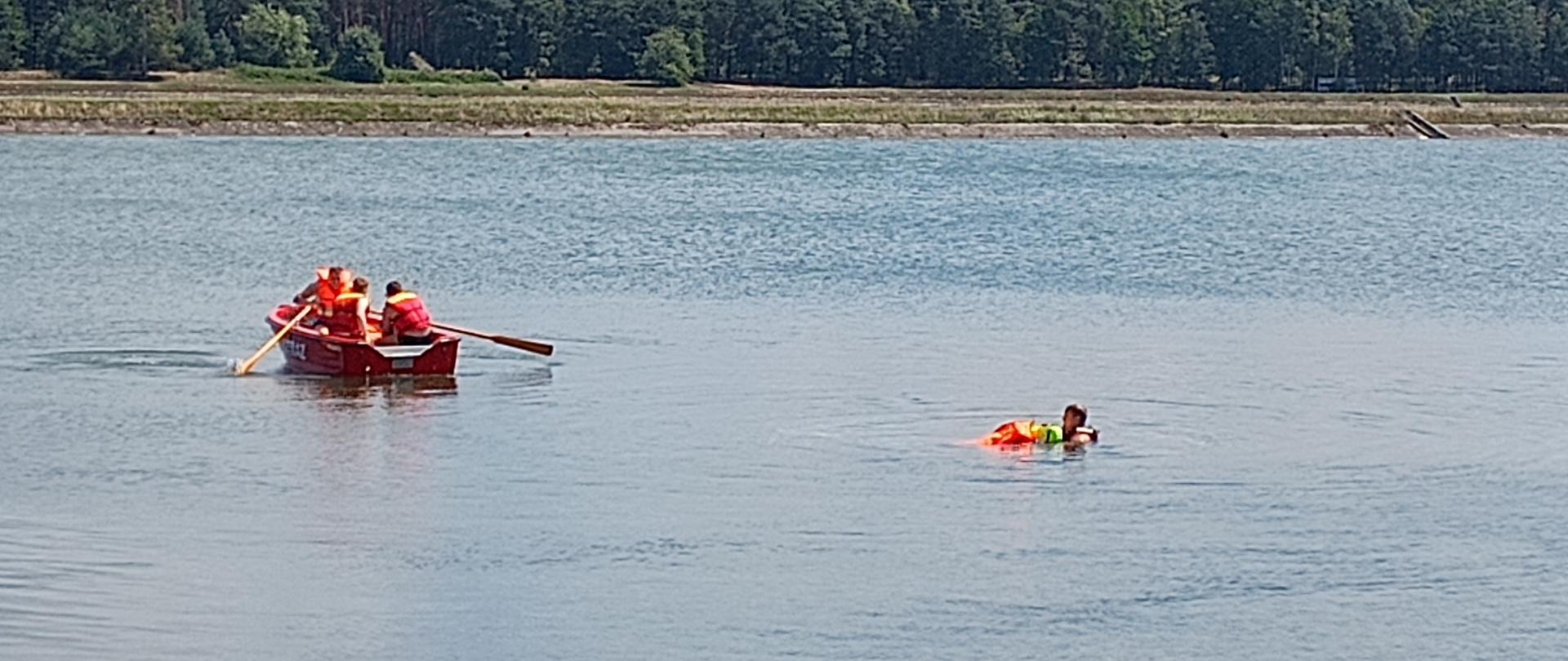 zdjęcie przedstawia akwen wodny na którym widzimy trzech strażaków w łodzi płynących w stronę poszkodowanego w tle las
