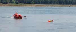 zdjęcie przedstawia akwen wodny na którym widzimy trzech strażaków w łodzi płynących w stronę poszkodowanego w tle las