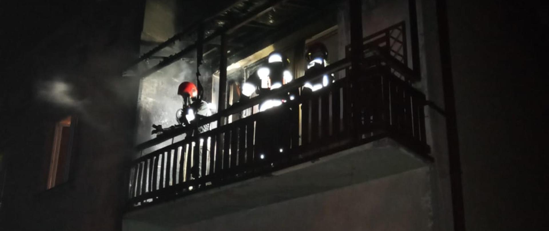 Zdjęcia przedstawia budynek wielorodzinny, na balkonie na pierwszym piętrze widoczni strażacy podczas dogaszania pożaru materiałów zgromadzonych na balkonie.