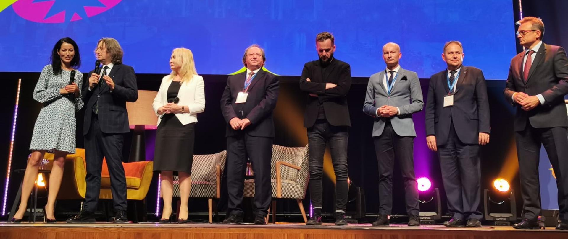 Duża grupa ludzi stoi na scenie, za nimi niebieski ekran z żółtym kodem QR.