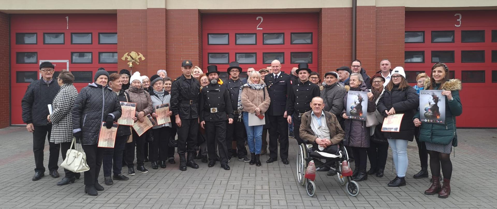 Przedstawiciele samorządu terytorialnego, zarządcy i administratorzy spółdzielni mieszkaniowych oraz seniorzy dziennego domu pobytu w Rypinie stojący na placu Komendy Powiatowej Państwowej Straży Pożarnej w Rypinie po odbytym spotkaniu z Zastępcą Komendanta Powiatowego Państwowej Straży Pożarnej w Rypinie i prewentystą Komendy Powiatowej Państwowej Straży Pożarnej w Rypinie. W tle znajdują się garaże jednostki ratowniczo-gaśniczej. 