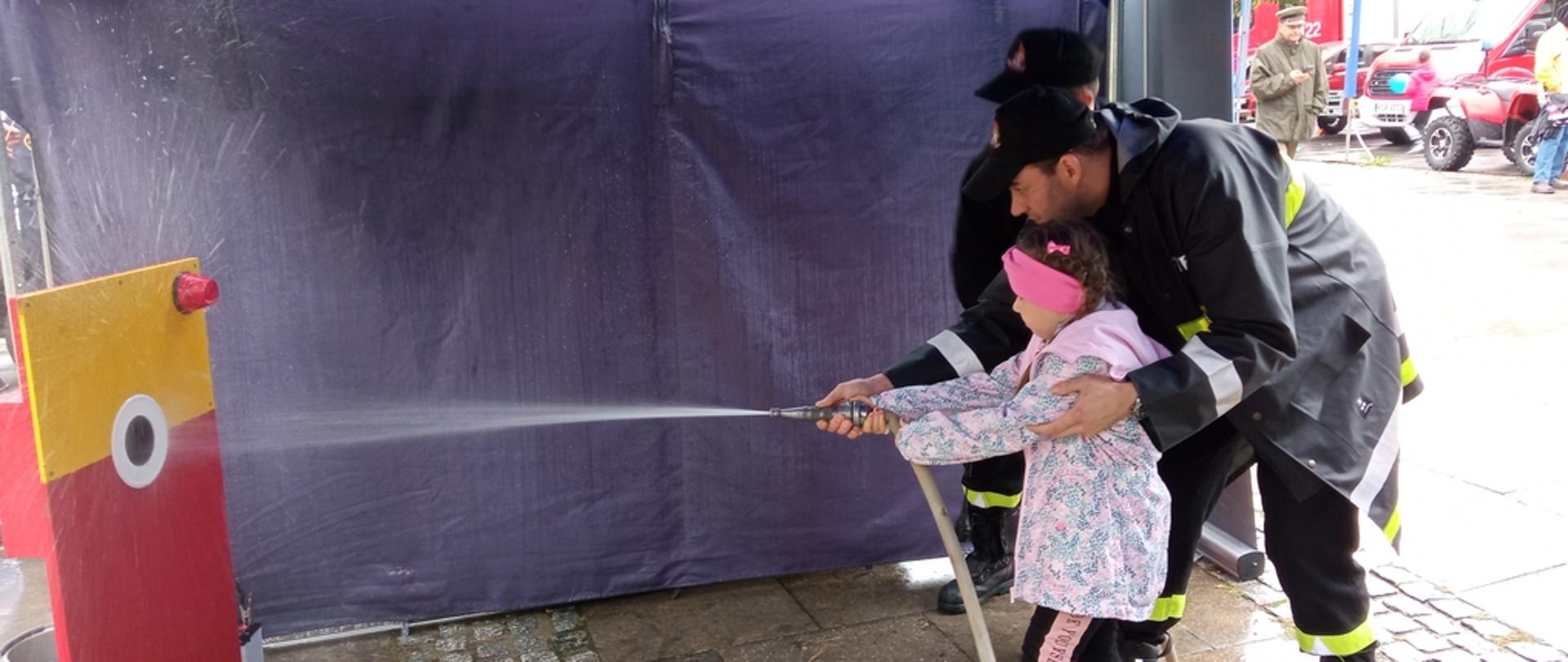 Na zdjęciu dziewczynka w jasnej kurtce z pomocą strażaka nalewa wodę do nalewaka przy użyciu hydronetki.