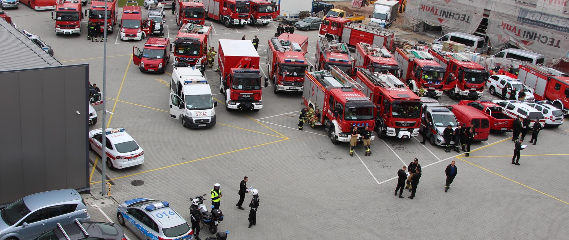 Zbiórka sił i środków na placu przy komendzie miejskiej w Kielcach. Na płycie stoją samochody pożarnicze przygotowane do wyjazdu. Z lewej strony pojazdy Policji eskortujące przejazdy kolumn aut na miejsce zdarzenia.
