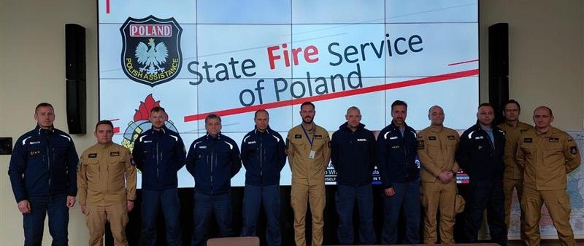  polscy i słowaccy strażacy stoją w szeregu pozując do zdjęcia, za nimi na monitorze wyświetlony napis State Fire Service od Poland