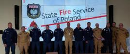  polscy i słowaccy strażacy stoją w szeregu pozując do zdjęcia, za nimi na monitorze wyświetlony napis State Fire Service od Poland