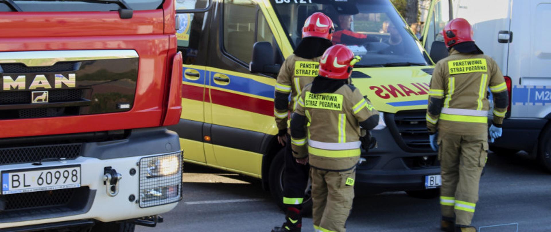 Widocznych jest trzech strażaków ubranych w ubrania specjalne. W tle znajdują się pojazdy uprzywilejowane: pojazd ratowniczo-gaśniczy oraz karetka. 