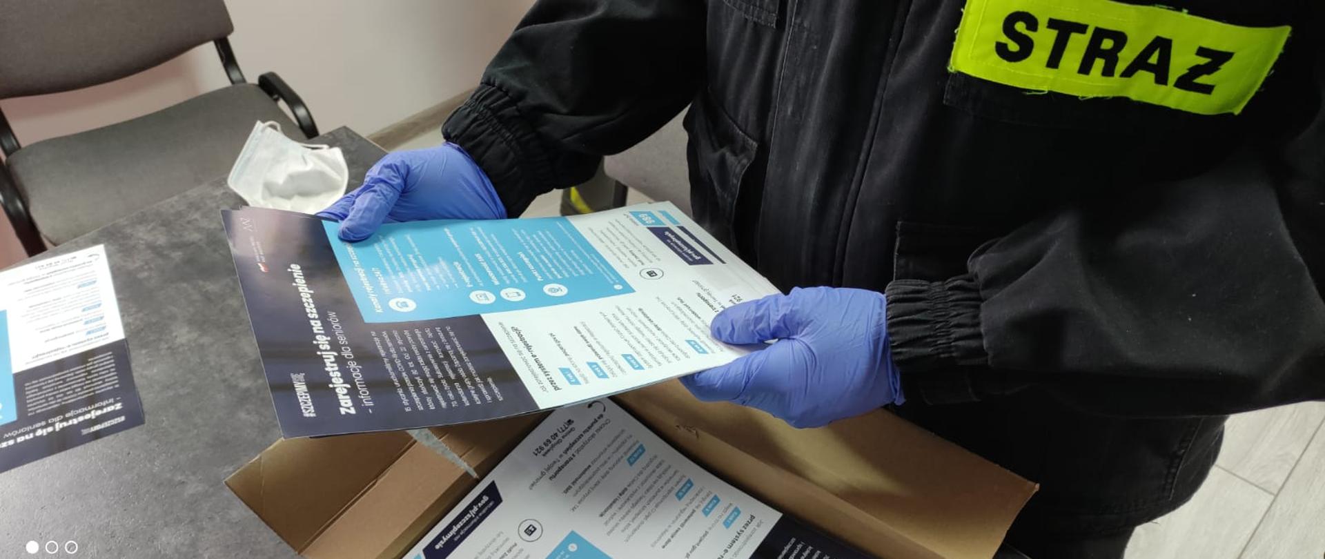 Zdjęcie przedstawia część tułowia strażaka OSP trzymającego w rękach ulotkę propagujące wiedzę na temat profilaktyki oraz szczepień przeciwko COVID-19.