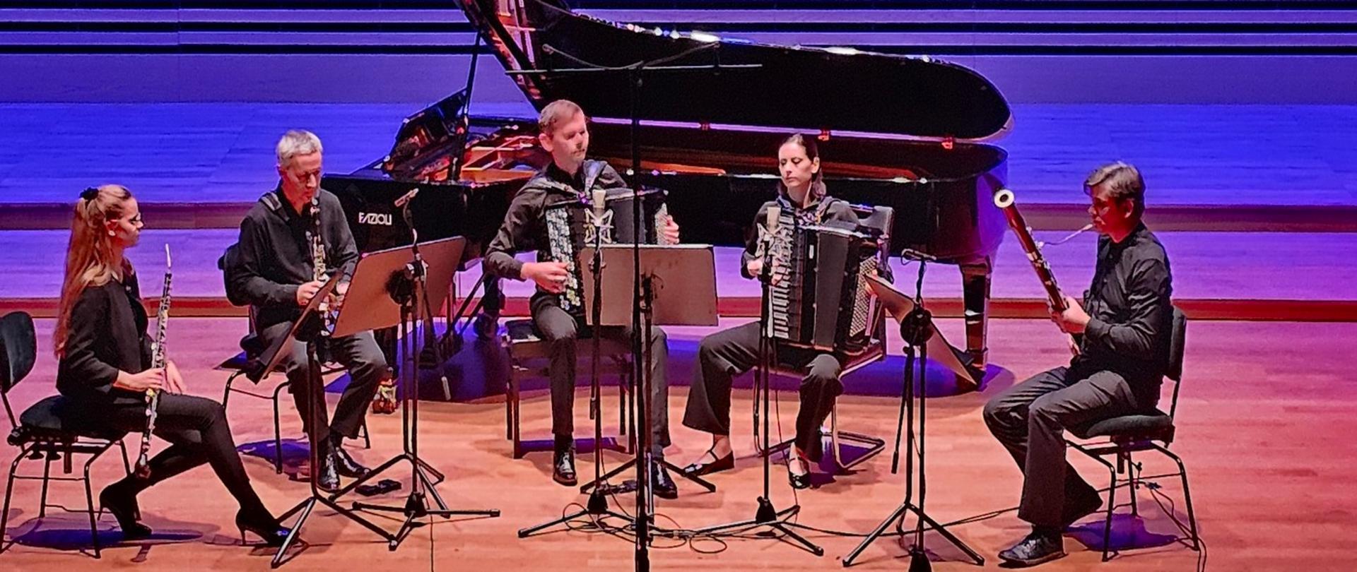 Dwie kobiety i trzech mężczyzn siedzi w półkolu na krzesłach grają na swoich instrumentach. Przed nimi stoją pulpity, za muzykami stoi fortepian.