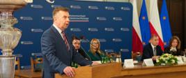 Minister Wieczorek stoi za mównicą obok stołu przy którym siedzi kilka osób, mówi do dwóch mikrofonów, za stołem granatowa ścianka z napisem Ministerstwo Nauki i Szkolnictwa Wyższego.