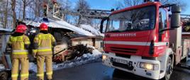 Na pierwszym planie po lewej stornie dwóch strażaków widocznych od tyłu, w tle budynek mieszkalny z zawalonym dachem, po prawej stronie wysunięta drabina mechaniczna nad dachem obiektu, w koszu drabiny stoi dwóch strażaków.