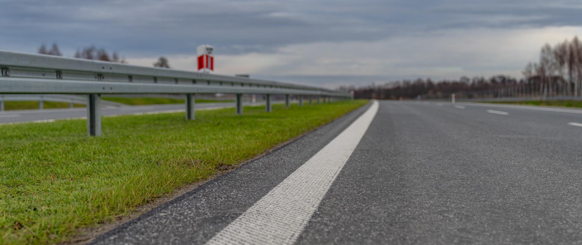 Widok na pas drogi ekspresowej z poziomu ziemi. W dużym zbliżeniu widać oznakowanie poziome, nawierzchnię i zieleń na pasie rozdziału.