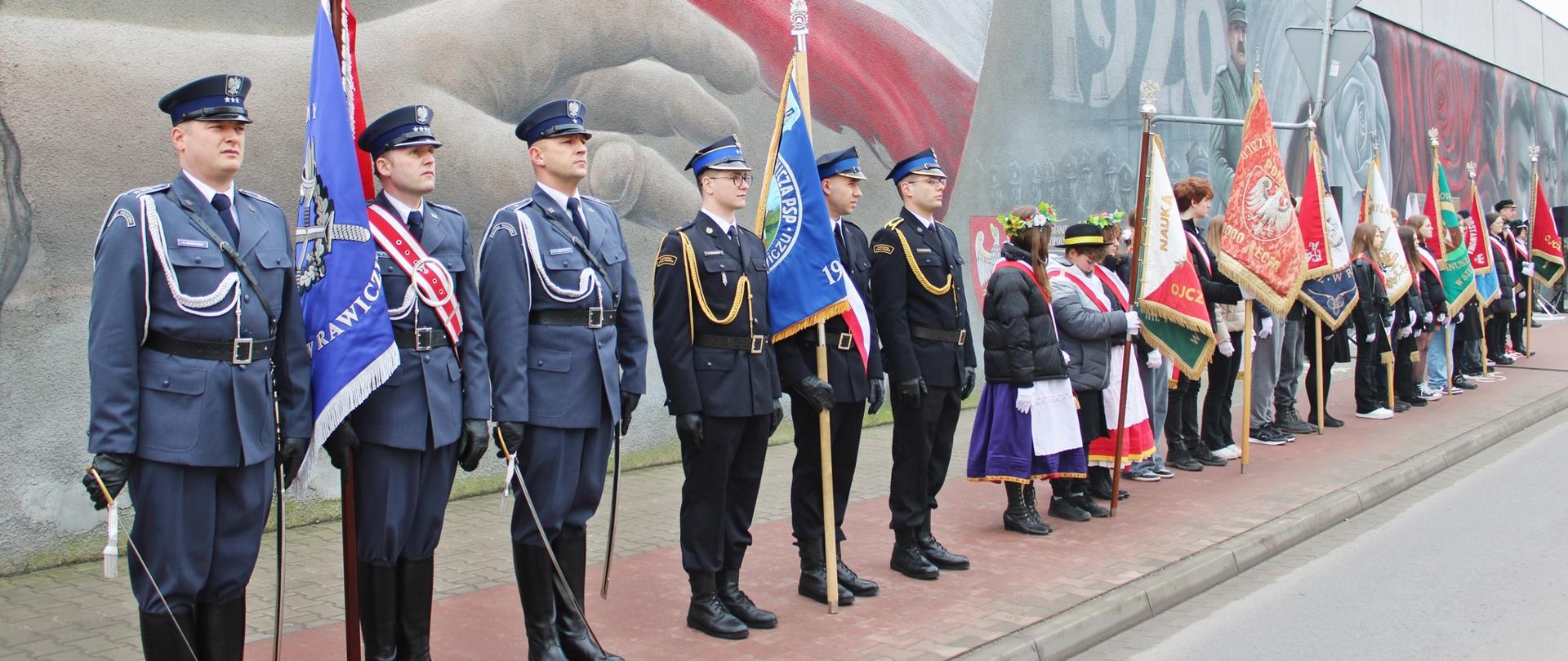Teren przed Zakładem Karnym w Rawiczu. Na tle muru więziennego, na którym znajduje się mural, na chodniku stoją w szeregu poczty sztandarowe. Pierwsze dwa od lewej strony to poczet ZK w Rawiczu oraz KP PSP w Rawiczu.