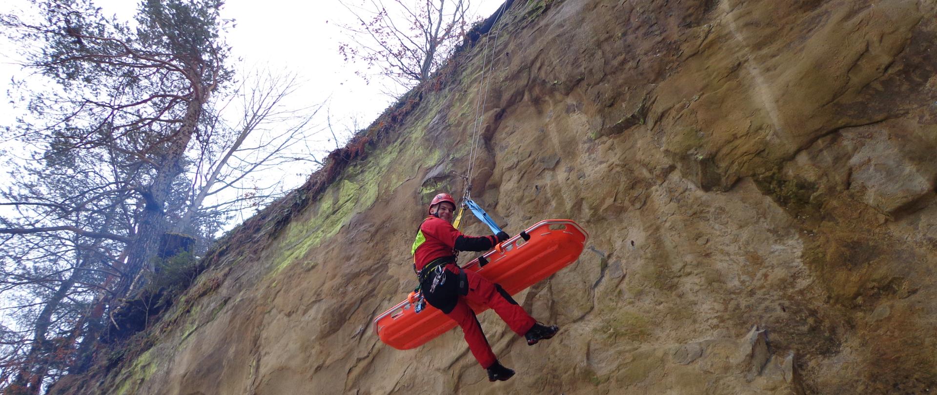 Zdjęcie przedstawia ratownika wysokościowego w czerwonym kombinezonie w uprzęży wysokościowej wpiętego w układ ratowniczy wraz z noszami. Ratownik porusza się po ścianie przeszkody naturalnej - kamienia. Stanowisko ratownicze zbudowane jest w oparciu o pewny punkt mocowania - drzewo.