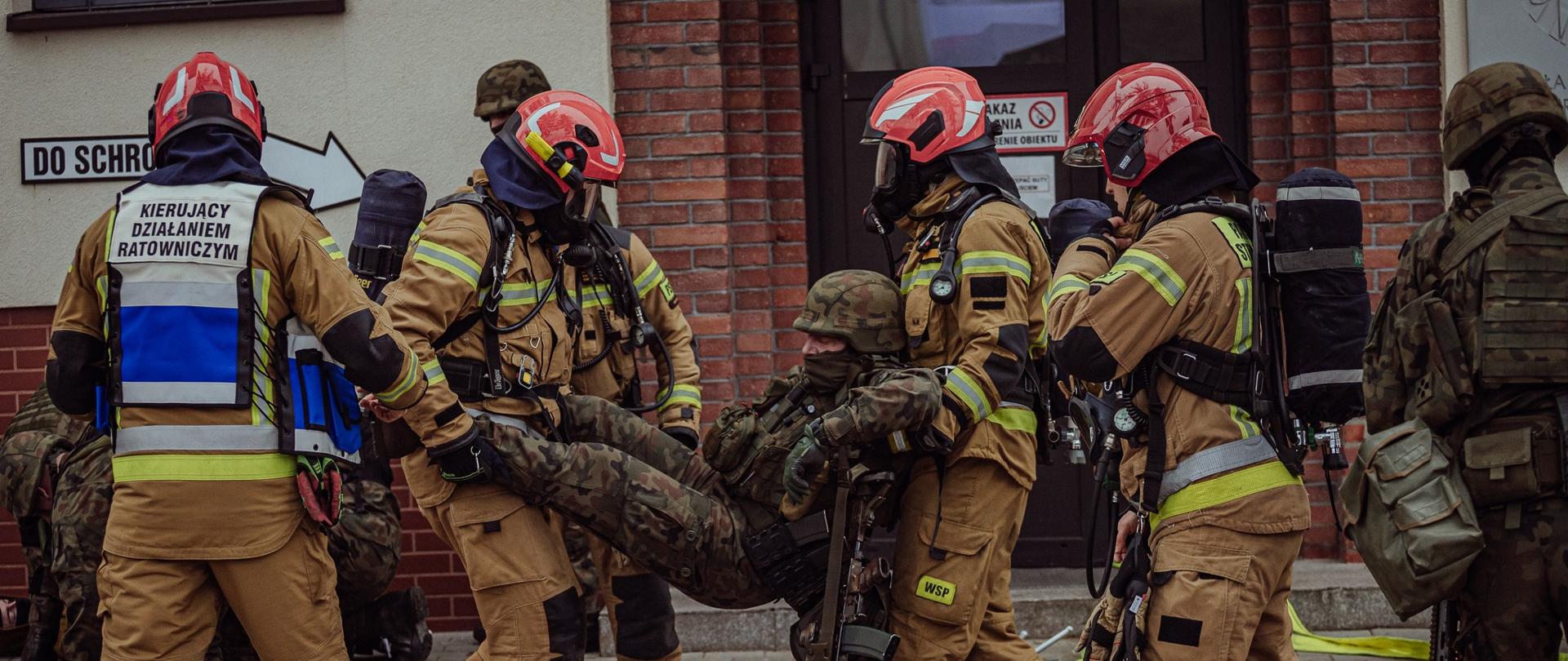 Strażacy w sprzęcie ochronnym przenoszą żołnierza w mundurze, ewakuując go sprzed budynku podczas ćwiczeń ratowniczo-wojskowych.