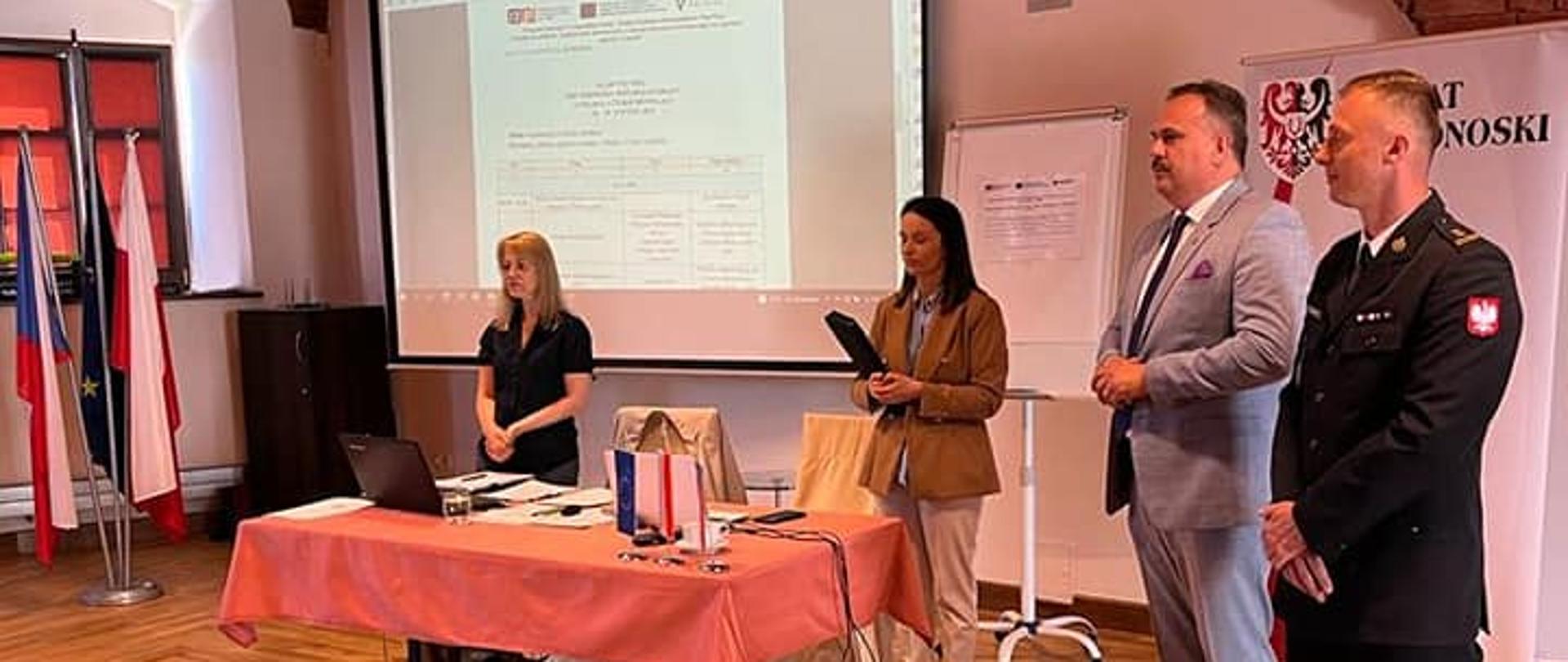 Na zdjęciu widać cztery osoby znajdujące się w pomieszczeniu dwie kobiety oraz dwóch mężczyzn. Jeden z nich to mężczyzna ubrany w czarne umundurowanie galowe straży pożarnej. Mężczyzna to strażak. Obok strażaka znajduje się Pan w garniturze. Panie znajdują się przy stole osłoniętym pomarańczowym obrusem. Jedna z Pań to naczelnik wydziału zarządzania kryzysowego, natomiast druga Pani to tłumacz. 