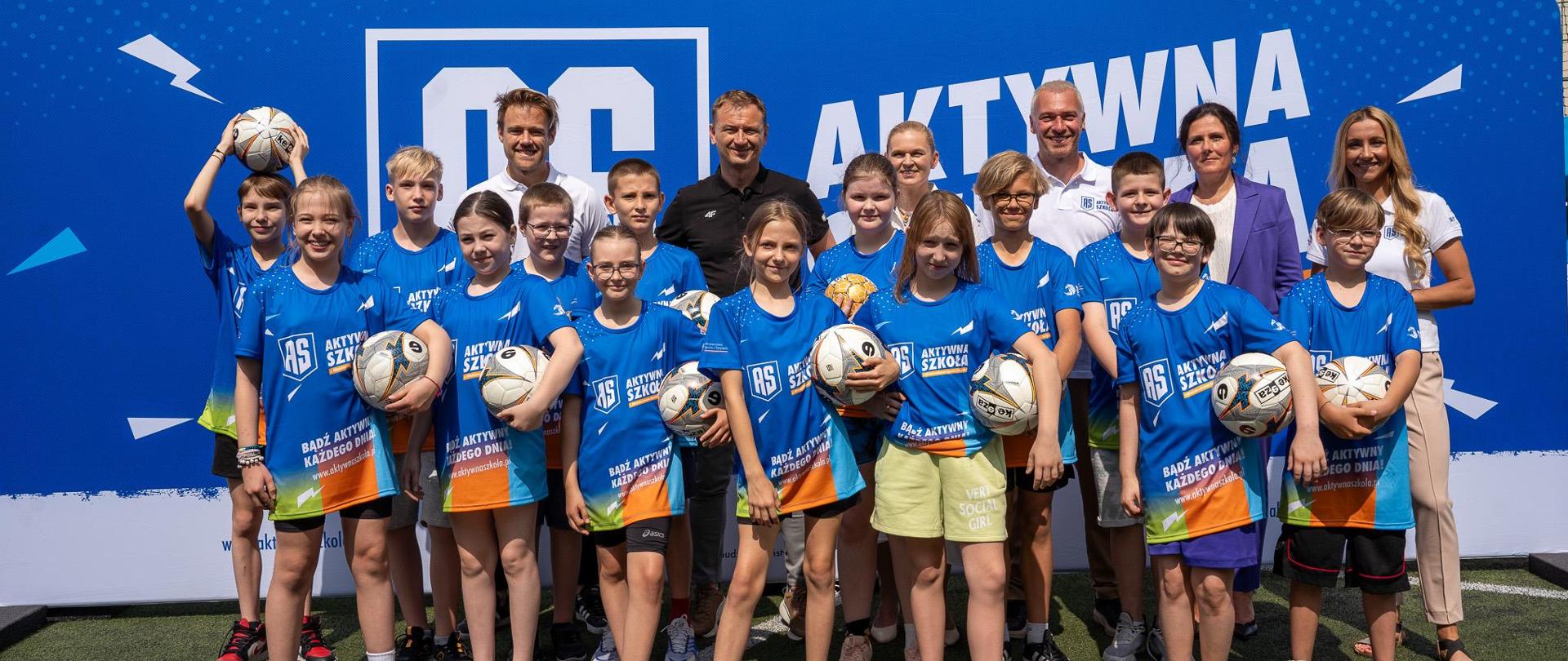 Zdjęcie zbiorowe. Na boisku sportowym na tle niebieskiej ścianki stoi grupa dzieci w niebieskich koszulkach z napisem Aktywna Szkoła, za nimi Ambasadorzy programu oraz minister Nowacka i minister Nitras.