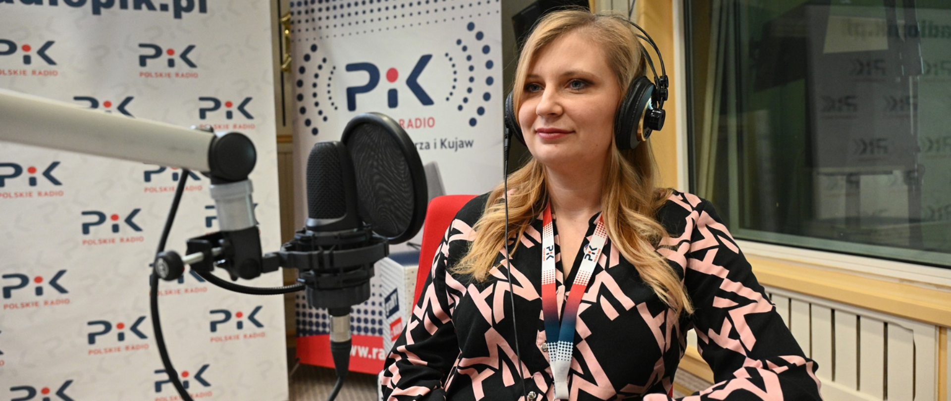 Kobieta w słuchawkach siedzi przy mikrofonie w studiu radiowym, przygotowana do nagrania audycji; w tle widoczne są banery z logo Polskiego Radia PiK.