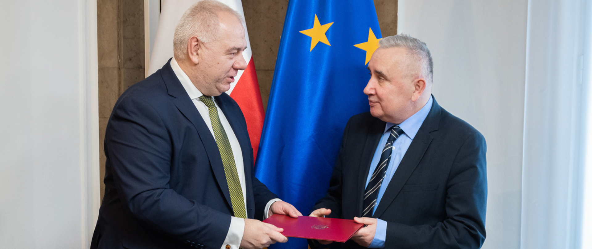Wicepremier Jacek Sasin wręcza wiceministrowi Piotrowi Pyzikowi akt powołania w bordowej teczce. W tle flagi polska i unijna