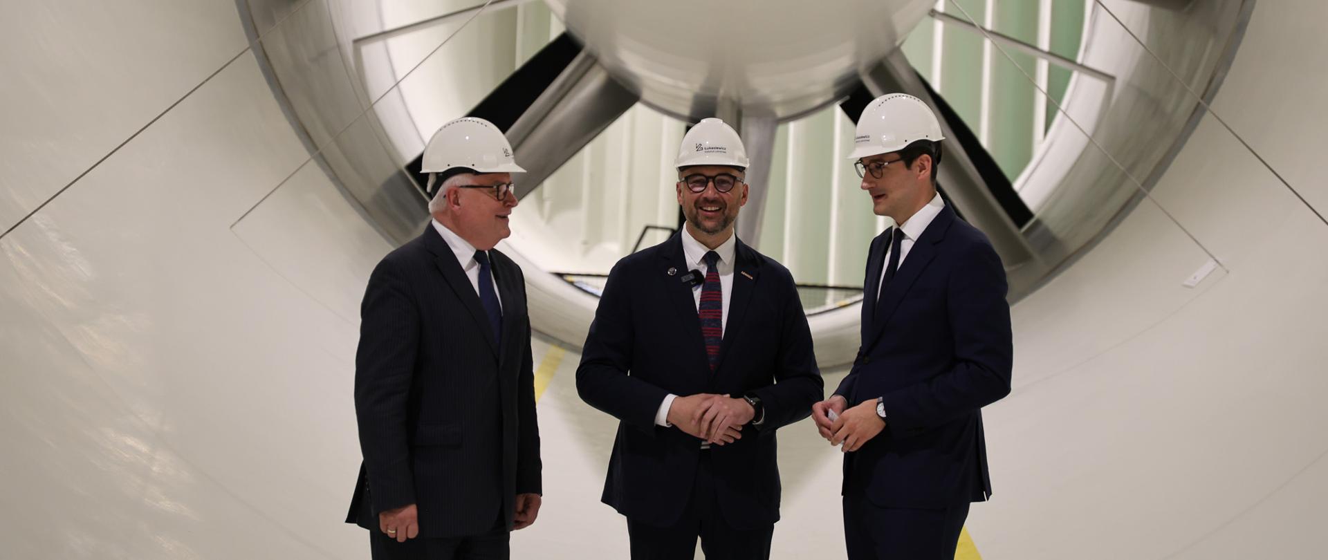 Wewnątrz białego tunelu stoi trzech mężczyzn w garniturach i białych kaskach, pośrodku minister Kulasek, za nimi wielka wielołopatowa turbina.