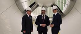 Wewnątrz białego tunelu stoi trzech mężczyzn w garniturach i białych kaskach, pośrodku minister Kulasek, za nimi wielka wielołopatowa turbin