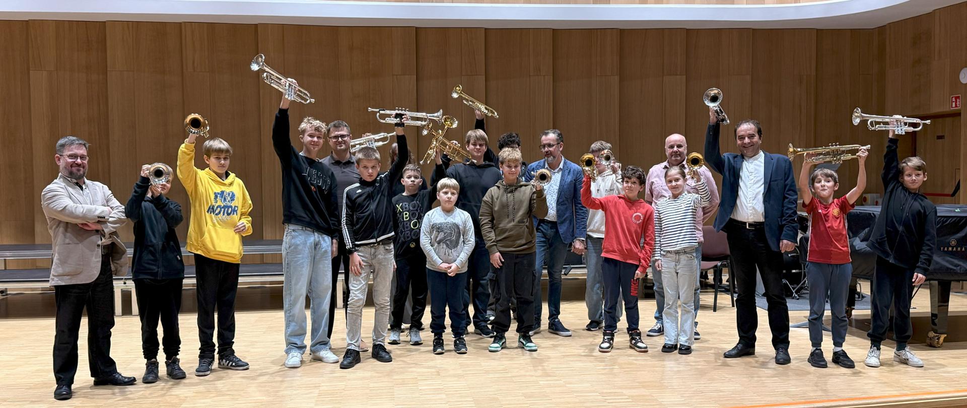 Uczniowie, nauczyciele, wykładowcy, scena. Uczniowie trzymają trąbki, miła, wesoła atmosfera. Fortepian, barwy ciepłe. Warsztaty muzyczne.