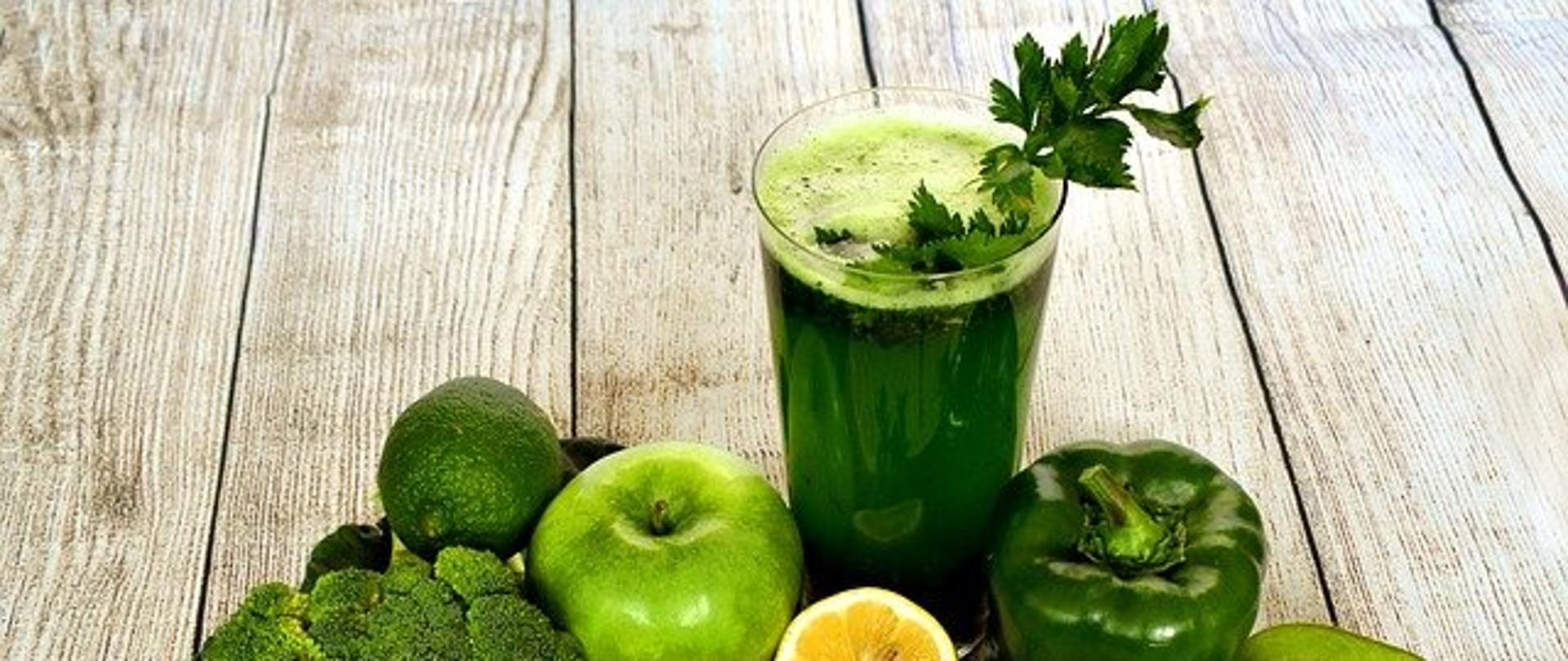 Na zdjęciu widoczna jest szklanka z zielonym sokiem oraz zielone warzywa i owoce w tym: jabłko, awokado, papryka, brokuł, kiwi, limonka i cytryna.