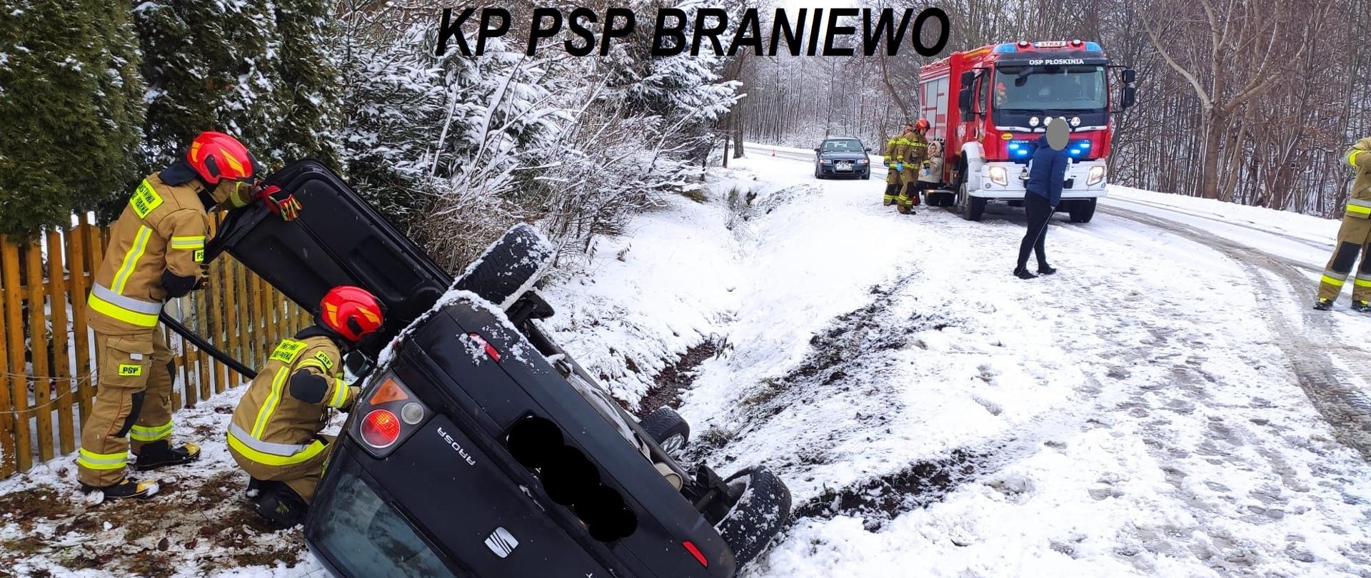 Ciemny samochód osobowy leży w rowie na dachu. Przy nim strażacy. Z prawej strony na drodze auto strażackie, dużo śniegu na drodze.