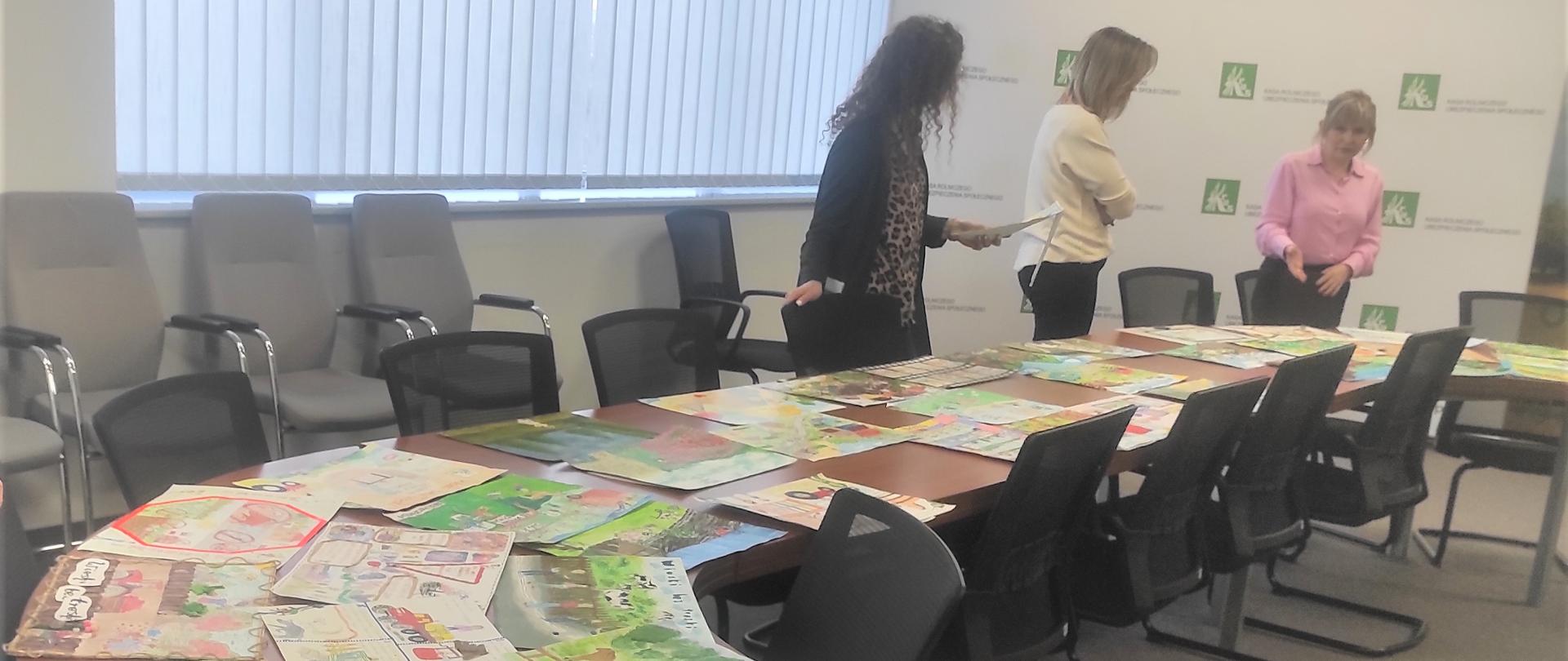Przy stole konferencyjnym stoją trzy kobiety i oglądają rozłożone na stole kolorowe prace plastyczne, przedstawiające m.in. traktory, dzieci, zwierzęta i krajobrazy wiejskie. W tle widoczna jest ścianka z logo KRUS.