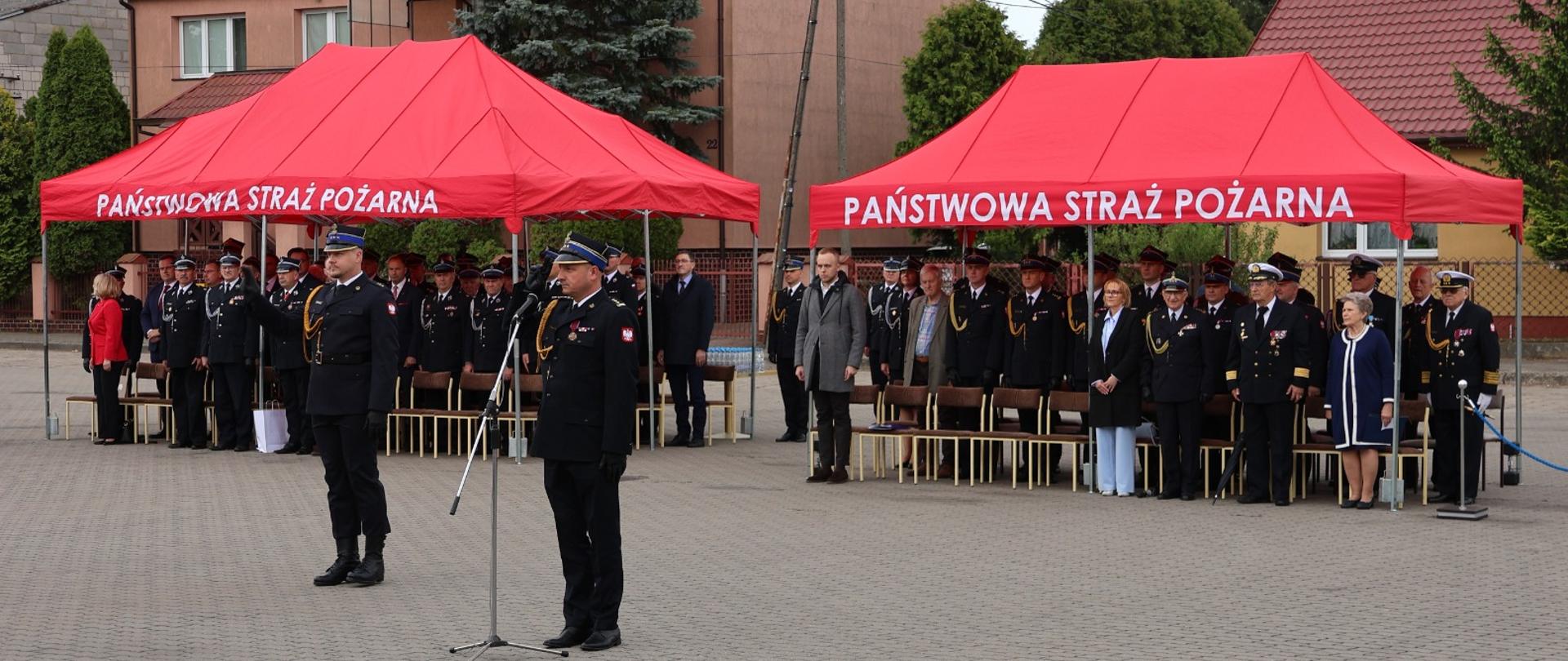 Uroczysty apel z okazji Dnia Strażaka na placu przed Komendą Miejską Państwowej Straży Pożarnej w Ostrołęce. Na pierwszym planie, przy mikrofonie, stoi Zastępca Komendanta Wojewódzkiego PSP w mundurze wyjściowym. W tle, pod dwoma dużymi czerwonymi namiotami z napisem „PAŃSTWOWA STRAŻ POŻARNA”, stoją zgromadzeni goście oraz funkcjonariusze w mundurach galowych. W tle widoczne są budynki mieszkalne oraz zieleń miejska.
