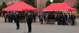 Uroczysty apel z okazji Dnia Strażaka na placu przed Komendą Miejską Państwowej Straży Pożarnej w Ostrołęce. Na pierwszym planie, przy mikrofonie, stoi Zastępca Komendanta Wojewódzkiego PSP w mundurze wyjściowym. W tle, pod dwoma dużymi czerwonymi namiotami z napisem „PAŃSTWOWA STRAŻ POŻARNA”, stoją zgromadzeni goście oraz funkcjonariusze w mundurach galowych. W tle widoczne są budynki mieszkalne oraz zieleń miejska.