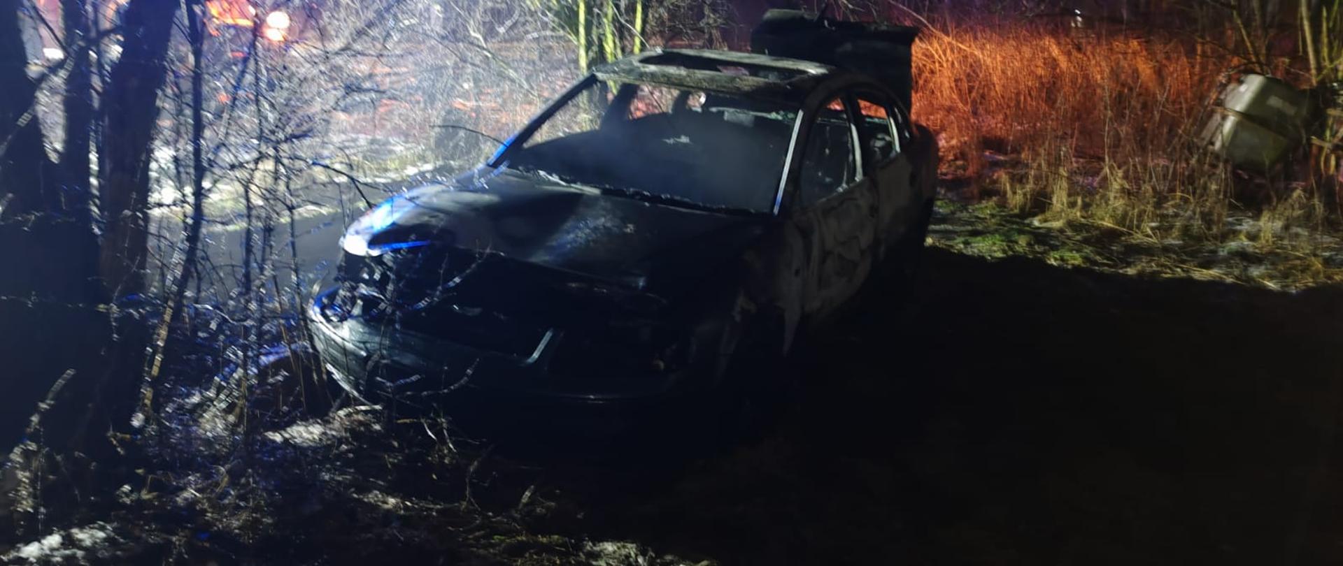 Nocne zdjęcie spalonego samochodu osobowego stojącego na poboczu w zaroślach; karoseria jest zwęglona, szyby zniszczone, a wokół widać osmaloną roślinność i wilgotne podłoże, z tyłu migoczą światła służb.