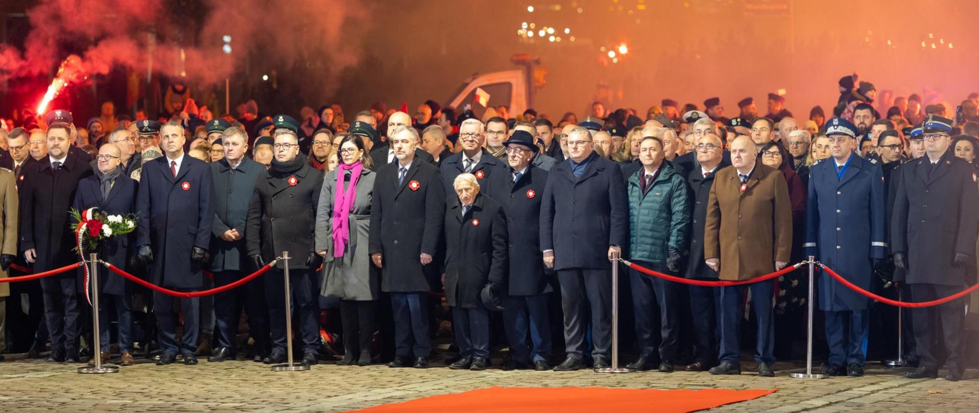 Zdjęcie przedstawia przedstawicieli władz i służb, zebranych gości podczas uroczystości. Wszyscy stoją za barierkami, w honorowym miejscu, na dworze.