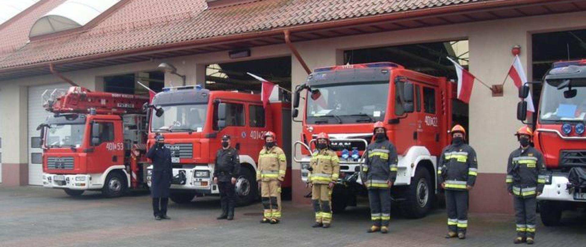 Zdjęcie przedstawiające pojazdy ratownicze komendy powiatowej w Busku-Zdroju. Przed pojazdami w kierunku flagi ustawiony Komendant Powiatowy, który salutuje. Przed pojazdami sześciu strażaków w pozycji "na baczność". Na ścianach budynku który jest w tle widoczne flagi państwowe.
