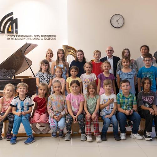 zdjęcie przedstawia nowych uczniów szkoły pierwszego stopnia we wspólnym ujęciu razem z dyrekcją w holu szkoły, w tle fortepian i logo szkoły na ścianie
